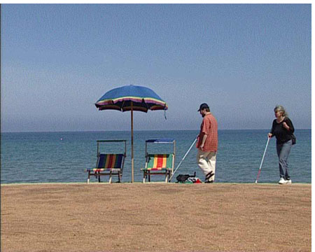 Friederike Kaivers und Joachim Steinbrück gehen mit Blindenstöcken am Meeresstrand auf Sonnenschirm und Liegestühle zu